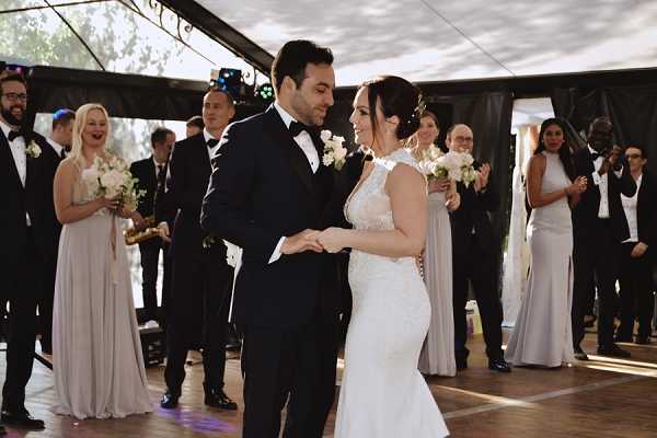 A couple shares their first dance under a large black and white marquee tent, with the groom in a black tuxedo and bow tie and the bride in a fitted lace wedding gown with a cap-sleeve neckline, holding a bouquet of white roses and greenery. The couple is looking down at their joined hands and smiling. Approximately 15–20 guests and bridal party members line the dance floor on either side, clapping and watching; bridesmaids wear floor-length light grey gowns and groomsmen wear black suits. The setting is an outdoor tented reception with visible stage lighting rigs and dappled natural light filtering through the tent edges, suggesting a daytime or late afternoon event.