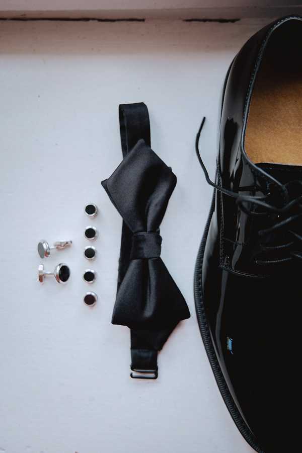 A flat lay detail shot of the groom's accessories arranged on a light grey surface, including one black patent leather lace-up Oxford shoe, a black satin pre-tied bow tie with an adjustable neck strap, a pair of silver cufflinks with black onyx or enamel insets, and four matching silver shirt studs with black centers. The accessories follow a classic black-and-silver formal color palette consistent with black tie attire. The composition is a close-up overhead flat lay with the shoe cropped at the right edge of the frame.