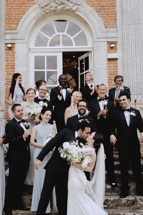 The couple shares a kiss at the base of the entrance steps of a grand brick chateau or manor house, surrounded by approximately twelve bridal party members and guests who are cheering, clapping, and catching confetti. The groom wears a black tuxedo with a bow tie, and the bride wears a white gown with a long veil, holding a large bouquet of white peonies and greenery. Bridesmaids are dressed in pale silver-grey halterneck gowns and carry smaller white floral bouquets, while the groomsmen wear black tuxedos with bow ties and white buttonhole flowers. The venue features a red brick facade with a white arched window above a double door entrance, through which a chandelier is visible, suggesting a classic formal French chateau setting. This is a wide group portrait shot taken outdoors on the steps.