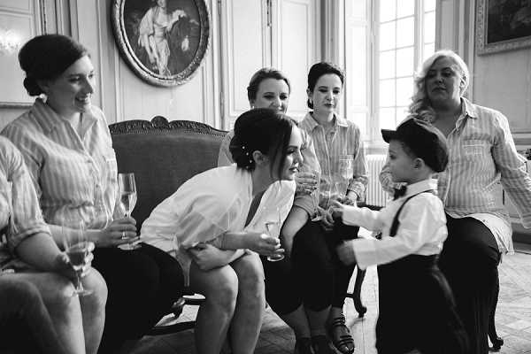 A black-and-white getting-ready scene inside what appears to be a chateau or manor house, featuring a bride and approximately four other women interacting with a young boy dressed in suspenders, dress trousers, and a flat cap. The bride, wearing a white button-down shirt with her hair pinned up, leans forward to engage with the child, who appears to be reaching toward her. Several women hold champagne flutes, suggesting a pre-ceremony celebration. The interior setting shows ornate molding, tall windows, a velvet sofa, and framed artwork on the walls, consistent with a classic French venue. The image is a candid mid-shot with warm contrast, capturing a relaxed, informal moment among the bridal party.