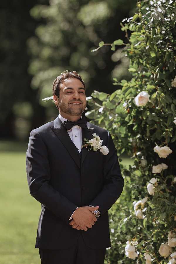 A portrait of the groom standing outdoors during the ceremony, positioned beside a tall floral arch or climbing rose structure covered in cream/white garden roses and green foliage. He is wearing a black tuxedo with a black bow tie and a white ranunculus boutonniere, and is smiling with his hands clasped in front of him. The setting appears to be a landscaped garden or chateau grounds, with a blurred green lawn and trees in the background. The shot is a medium portrait framing the groom from approximately the waist up.