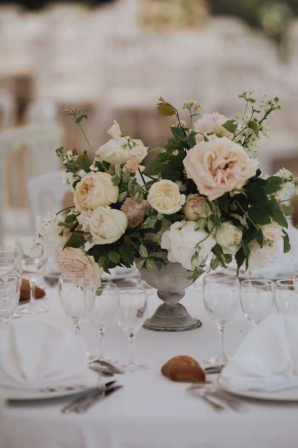 Close-up detail shot of a wedding reception table centerpiece arranged in a weathered stone-effect urn pedestal vase. The floral arrangement features blush garden roses, cream and ivory peonies, white ranunculus, and small white filler flowers with abundant green foliage. The table is set with a white linen tablecloth, white folded napkins, silver cutlery, crystal wine glasses, and small bread rolls placed directly on the cloth. The background shows a softly blurred reception setting with additional white-covered chairs and tables, suggesting an outdoor or marquee venue. The overall decor palette is blush, cream, and ivory with a classic, romantic styling.