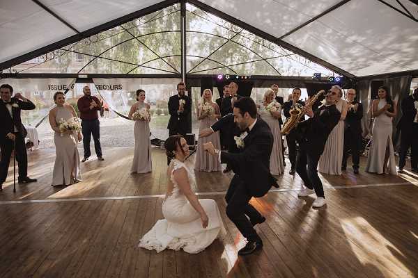 The bride and groom are performing a lively, choreographed first dance inside a clear-top marquee tent with a polished wooden dance floor. The groom, wearing a dark navy suit with a white boutonniere, is crouching low while the bride, in a fitted white lace gown, appears to be sitting or dropping to the floor in a playful dance move. A live band is performing behind them, including a saxophonist and trumpet player, while the bridal party — bridesmaids in floor-length light taupe/champagne gowns holding white bouquets and groomsmen in black tuxedos — lines the perimeter of the dance floor cheering and clapping. The image is a wide shot capturing the full scene, with natural light filtering through the transparent marquee roof.