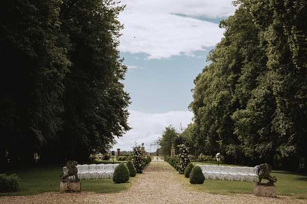 An outdoor wedding ceremony setup photographed from a wide, symmetrical perspective down a long gravel tree-lined avenue. Two rows of white chairs are arranged on either side of a central aisle, flanked by neatly trimmed box hedges and stone or bronze sculptural figures at the entrance. Clusters of white floral arrangements are positioned along the aisle at intervals. The ceremony space is set within formal French garden grounds featuring tall mature trees on both sides, with iron gates visible in the distance at the far end of the avenue. No guests or wedding party are present, suggesting the image was taken before the ceremony. Potential venue feature image.