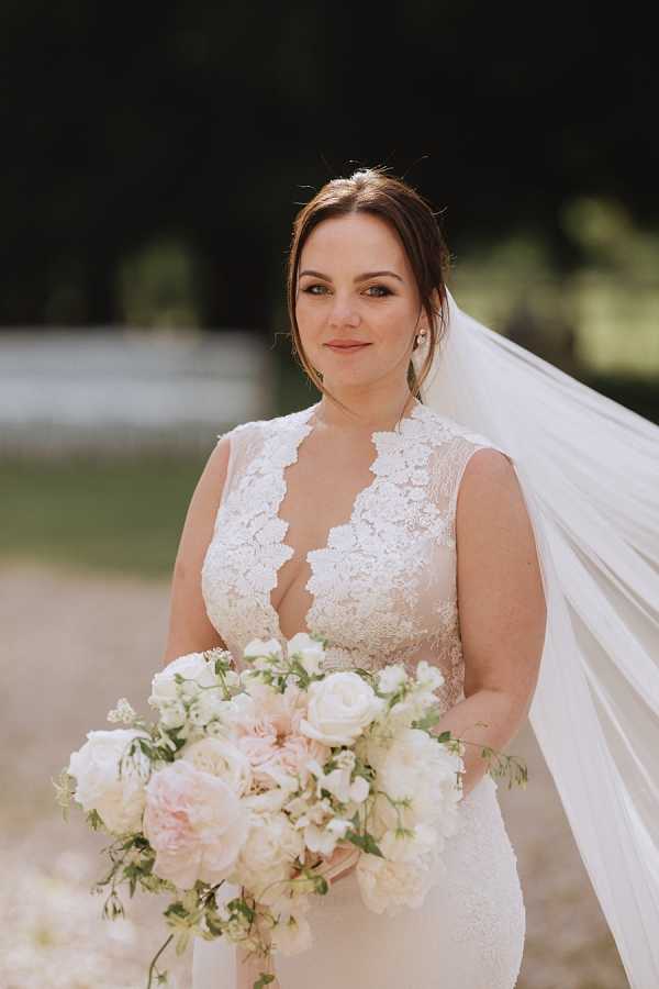A bridal portrait taken outdoors, showing the bride from approximately the waist up against a softly blurred background of trees and a gravel path. The bride wears a sleeveless fitted gown with a deep V-neckline edged in lace appliqué detailing and a long cathedral-length veil that trails to one side. Her dark brown hair is styled in an upswept chignon, and she wears small stud earrings. She holds a loose, cascading bouquet composed of blush garden roses, white ranunculus, white sweet peas, and trailing greenery. The overall color palette is soft blush and white. The shot is a medium portrait with a shallow depth of field, taken in natural daylight.