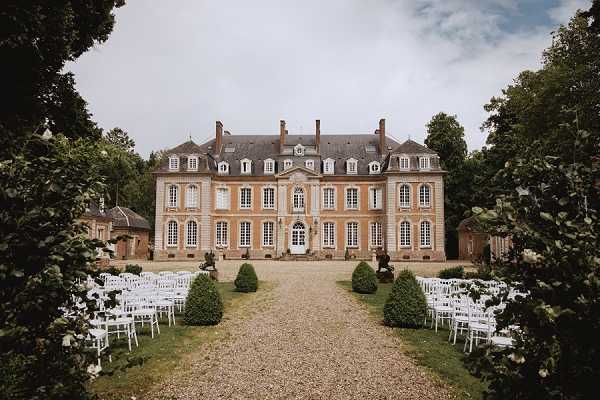 An outdoor wedding ceremony setup in front of a classic French chateau with warm ochre-toned stone facade, mansard roof, and symmetrical architecture. Rows of white chairs are arranged in two sections along a gravel pathway leading up to the chateau entrance, set up for a ceremony with no guests yet present. The seating is framed by manicured cone-shaped topiary shrubs on either side of the central path. Wide establishing shot capturing the full facade and ceremony layout. Potential venue feature image.