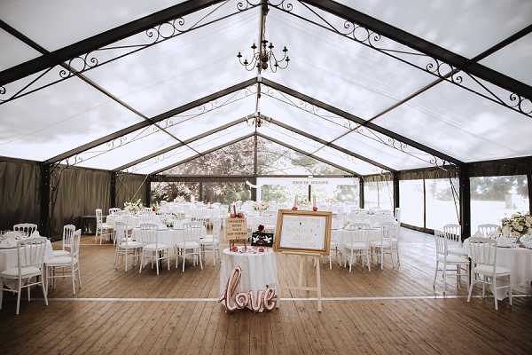 A wide shot of a wedding reception setup inside a large clear-roof marquee tent with black iron frame detailing and a wrought-iron chandelier overhead. The tent features transparent walls and roof panels that allow natural light to fill the space, with wooden plank flooring throughout. Round tables dressed in white linens are arranged around a central dance floor, surrounded by white Chiavari chairs, with white floral centerpieces visible on the tables. In the foreground, a small welcome table holds a gold-framed seating chart on an easel, a light-up rose gold 'love' sign, and additional decor items, establishing a white and rose gold color palette for the event. Potential venue feature image.