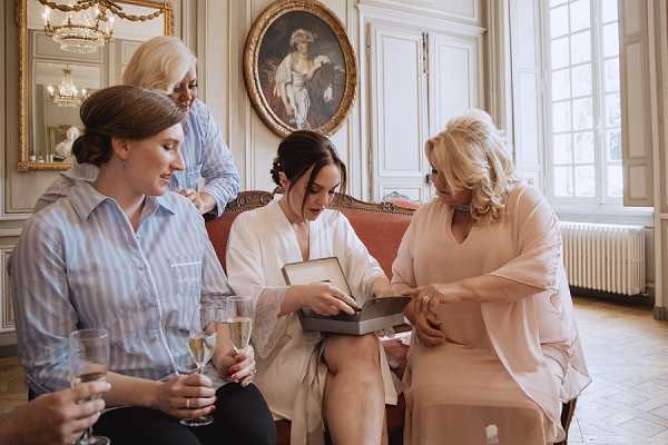 A getting-ready scene taking place inside a chateau or manor house, featuring four women gathered around a pink velvet sofa in a ornate room with white boiserie paneling, a gilded mirror, and a classical oil painting on the wall. The bride, wearing a white robe, sits at the center opening a gift box that appears to contain jewelry, while a woman in a blush pink wrap top leans in to look; a third woman in a light blue striped shirt holds a champagne flute and watches, and a fourth woman standing behind assists with something at her neckline. The setting features warm natural light from a large window and the room's classic French interior decor creates a formal, traditional atmosphere. Medium shot capturing all four figures in a candid, intimate moment.