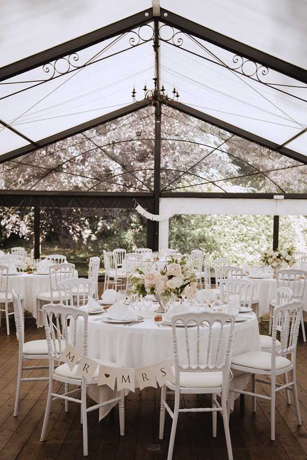 A reception setup inside a clear-roofed marquee tent with black metal frame detailing and a small chandelier hanging from the apex. The tent features transparent panels on all sides, allowing views of surrounding trees. Round tables are dressed in white floor-length linens and set with glassware and white folded napkins, surrounded by white Napoléon chiavari chairs. The foreground table is identified as the sweetheart table with a kraft paper "MR & MRS" bunting banner draped across two chairs. Centerpieces consist of blush pink and ivory floral arrangements including what appear to be garden roses and ranunculus with greenery. The overall decor palette is white and blush with a classic, romantic theme. Wide interior shot taken from a standing angle showing multiple guest tables receding into the background.