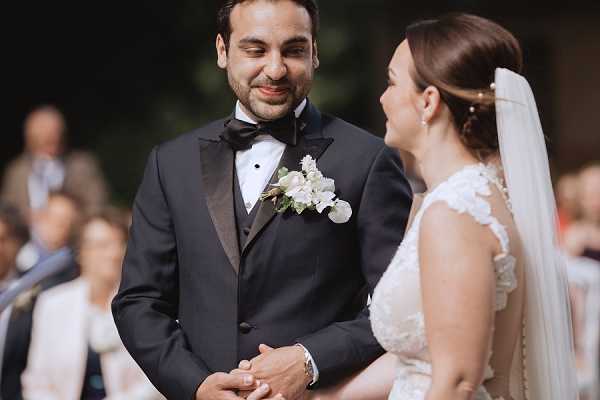 A close-up portrait of a couple during an outdoor wedding ceremony, with the groom smiling downward toward the bride as they hold hands. The groom wears a black tuxedo with satin lapels, black bow tie, and a white floral boutonniere with small greenery. The bride wears a lace cap-sleeve wedding gown and a cathedral-length veil, with her hair styled in an updo. Seated guests are visible in soft focus in the background, suggesting a formal outdoor ceremony setting. The overall styling is classic and formal.
