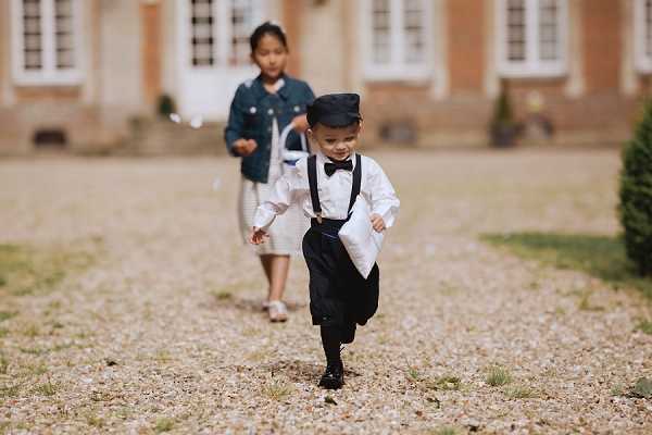 Two children are running along a gravel path in front of a French chateau-style building during what appears to be a wedding celebration. In the foreground, a young boy dressed in a white dress shirt, black suspenders, black shorts, black socks, and a flat cap is running toward the camera. Behind him, a girl wearing a denim jacket over a light-colored dress follows along the path. The shot is a medium portrait taken at ground level, with the château facade softly blurred in the background.