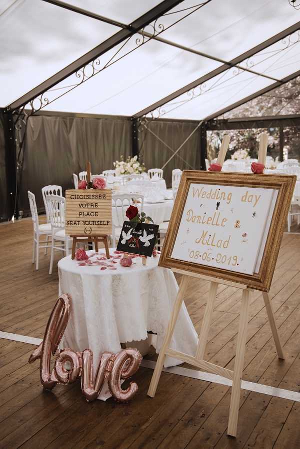 Detail shot of a wedding reception entrance display set up inside a clear-roofed marquee tent with dark fabric side panels and a wooden floor. A gold-framed welcome sign on a wooden easel reads 'Wedding day Danielle & Milad 08.06.2019' in rose gold lettering, positioned next to a small round table draped in a white linen cloth. The table holds a wooden letter board sign reading 'Choisissez votre place / Seat yourself,' a small chalkboard with dove illustrations, and scattered red rose petals and pink roses. A rose gold foil 'love' balloon script sits on the floor in front of the table. In the background, round banquet tables are set with white linens and white Napoleon chairs, with white floral centerpieces visible, indicating a classic French reception style with a rose gold and red color palette.