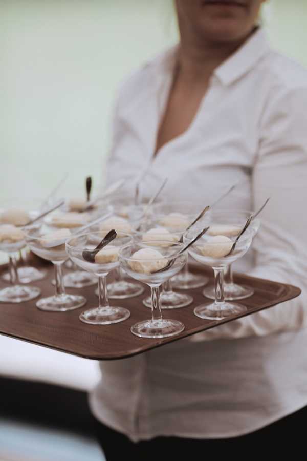 Close-up shot of a server in a white button-down shirt holding a rectangular brown tray loaded with approximately a dozen coupe-style glass dessert dishes, each containing what appears to be a pale cream or vanilla-colored sorbet or mousse scoop with a small silver spoon resting inside. The tray is being carried at waist level against a softly blurred neutral background. The styling is clean and classic, consistent with a formal wedding reception or cocktail hour service.