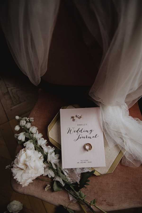A getting-ready flat lay detail shot arranged on a dusty rose velvet chair seat. The central item is a blush pink personalized wedding journal titled 'Danielle's Wedding Journal – The Future Mr. and Mrs. Jokar,' placed on a gold hexagonal tray. Resting on the journal cover are a gold wedding band and small crystal or diamond stud earrings. To the left of the tray, a loose arrangement of white flowers includes ivory peonies, white delphinium stems, and small white buds with green foliage. The edge of a white tulle veil drapes across the upper portion of the frame. The overall decor palette is blush, gold, and white, consistent with a classic or modern romantic styling theme.
