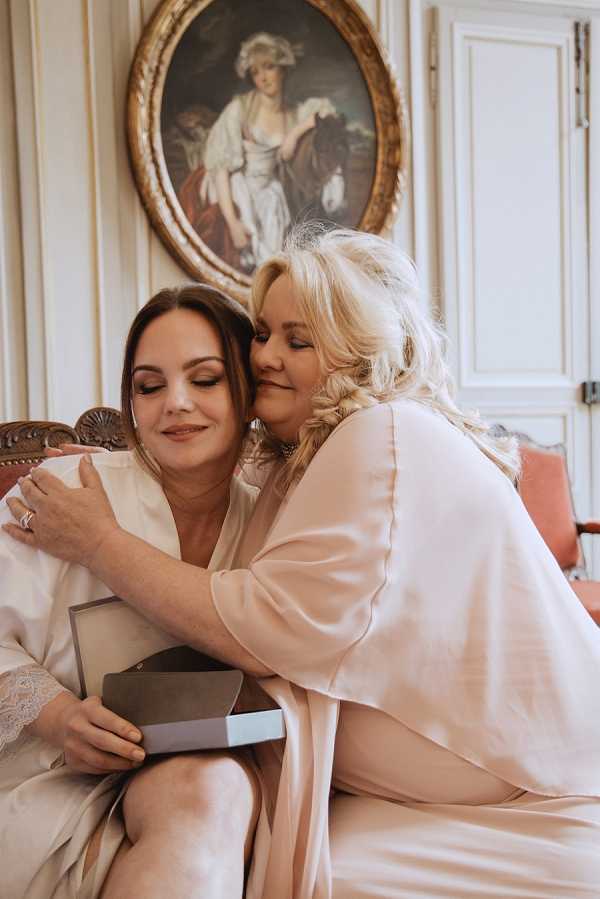 A getting-ready moment captured indoors, showing two women seated together on an ornate carved wooden sofa with coral/salmon upholstery. The bride, wearing a cream satin robe with lace trim, holds an open gray gift box while the other woman — likely her mother — wearing a blush champagne satin robe leans in to kiss her cheek in an embrace. Both have their eyes closed and are smiling softly. A small ring is visible on the bride's hand. The room features classic French interior decor with white wood paneling and a large oval gilt-framed portrait painting of a woman on the wall behind them. The shot is a close-up portrait with warm, soft natural lighting.