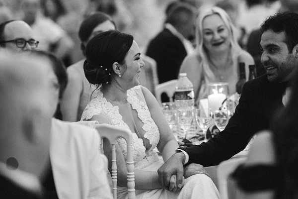 A black-and-white candid shot taken during the wedding reception, capturing the bride and groom seated at the head table holding hands and laughing together. The bride wears a lace-detailed gown with a V-neckline and has her dark hair styled in an updo with small hair accessories. The groom, in a dark suit, leans toward her with a broad smile. Behind them, guests seated at surrounding tables are also laughing, with glassware and candles visible on the tables. The image is a medium close-up portrait with soft contrast and a shallow depth of field that keeps the couple sharp while the background guests are softly blurred.