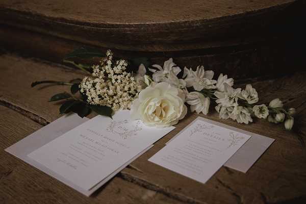 A close-up flat lay detail shot of wedding stationery arranged on a rustic wooden surface. Two printed cards — appearing to be an invitation and a details card — feature delicate botanical line illustrations and script typography in a neutral, muted palette with light grey or warm white card stock. Alongside the stationery, a small arrangement of cream and white florals is loosely scattered, including a garden rose, white stock flowers, small white clusters resembling elderflower or astilbe, and green foliage. The overall styling is minimal and classic, with a neutral, soft tone throughout.