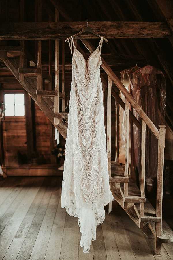 A detail shot of a wedding dress hanging from a wooden beam inside a rustic barn setting. The gown is a fitted, spaghetti-strap slip-style dress with intricate all-over lace featuring a bold, geometric floral pattern in ivory over a nude/champagne lining, giving it a boho aesthetic. The dress hangs beside a weathered wooden staircase with exposed timber framing, wide-plank floors, and warm dark wood tones throughout. A second garment in a plastic dry-cleaning bag is visible in the background to the right.