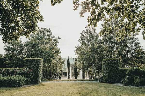 Wide shot of a formal garden pathway leading toward a glass or mirrored structure in the background, framed by tall clipped hedges on either side and mature trees overhead. The grounds feature a manicured lawn and cypress trees flanking the central axis, creating a symmetrical approach to what appears to be a contemporary pavilion or orangerie-style building. No people are visible in the frame. The image has a soft, slightly overexposed tone with muted greens. Potential venue feature image.