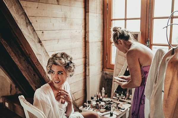 A bride is getting ready in a rustic wooden-walled room, seated at a vanity table covered with makeup products and beauty tools while wearing a white lace robe. She has an upswept blonde hairstyle with soft waves and is smiling toward the camera. A second woman, likely a bridesmaid or makeup artist, stands beside her wearing a strapless purple/mauve dress and appears to be reviewing something on a phone or device. The warm, natural light comes through a wooden-framed window, and a garment bag is visible hanging in the background. The setting has a rustic, barn-style aesthetic, and the image is a medium portrait shot with a slightly warm color grade.
