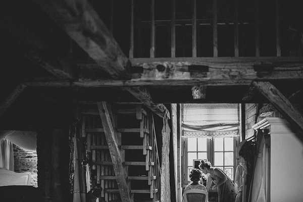 A black-and-white getting-ready scene set inside a rustic barn or farmhouse interior, featuring exposed wooden beams, a steep wooden staircase, and a multi-pane window that provides the primary light source. Two figures — likely the bride and a hair or makeup artist — are positioned near the bright window in the background, one seated in a chair while the other attends to her. The wide interior shot uses deep contrast between the dark, heavy timber structure in the foreground and the bright window light illuminating the two women, creating a dramatic chiaroscuro effect. The setting has a rustic, unfinished aesthetic with raw wooden construction throughout.