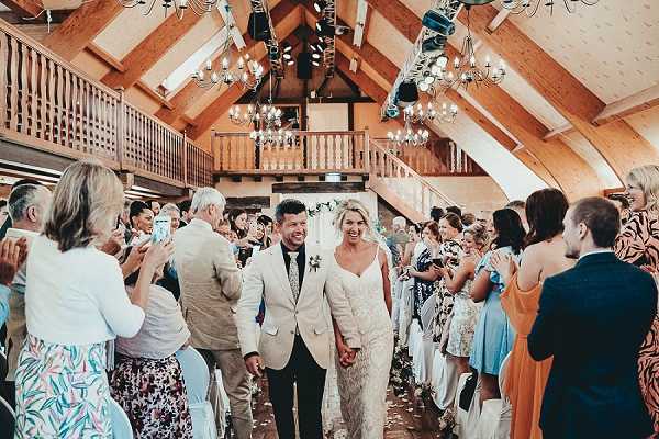 The bride and groom walk back up the aisle together immediately after their ceremony inside a barn venue with exposed wooden beams, a vaulted ceiling, and a mezzanine balcony level above. The groom wears a beige/cream blazer with dark trousers and a black tie, while the bride wears a fitted lace gown with spaghetti straps. Approximately 50 or more guests line both sides of the aisle, clapping and photographing the couple on smartphones as they pass. The barn is lit by two small crystal chandeliers and additional stage-style lighting mounted on the ceiling beams. The wide-angle shot captures the full length of the aisle with confetti scattered on the floor, and a floral arch is faintly visible at the altar in the background.