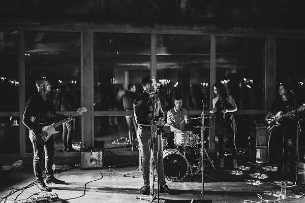 Black-and-white image of a live band performing at a wedding reception. The band consists of approximately five to six musicians including a lead vocalist at the microphone, two guitarists, a drummer, and at least one additional player, all dressed in dark clothing. The performance takes place on a hard floor in front of large glass sliding doors or windows, through which guests can be seen socializing in a warmly lit interior space. Stage monitors, amplifiers, cables, and drum kit hardware are visible on the floor. The shot is taken from a slightly low wide angle, capturing the full band setup and the venue interior in the background, with deep shadows and high contrast typical of low-light black-and-white photography.