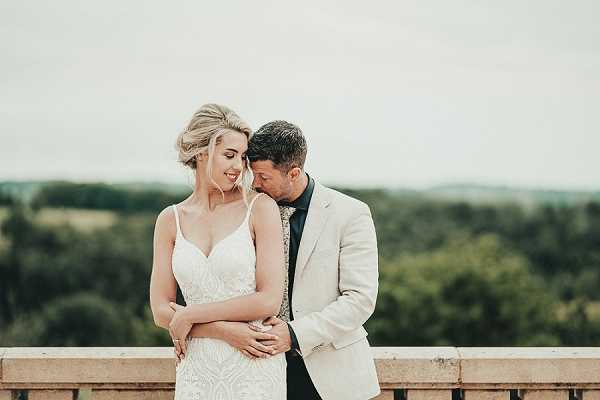 A couple portrait taken outdoors on a stone balustrade terrace, with a wide countryside view visible in the background. The bride wears a fitted white lace spaghetti-strap gown with a V-neckline and has her blonde hair styled in an updo, while the groom wears a beige/champagne suit jacket with dark trousers and a gold-toned tie. The groom stands behind the bride, leaning his head close to hers in an intimate pose, with his arms wrapped around her waist. The shot is a medium portrait composition with a soft, slightly hazy natural light giving the image a warm, muted tone.