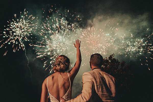 A couple watches a fireworks display during their wedding reception, photographed from behind in a wide portrait shot at night. The bride wears a low-back white gown with lace detailing and spaghetti straps, her hair styled in an updo, while the groom wears a light beige or tan suit jacket. The groom has his arm around the bride, and one of them raises a hand upward toward the sky. Multiple large fireworks burst in shades of gold, white, and teal-green against a dark night sky, creating a dramatic backdrop to the silhouetted couple.