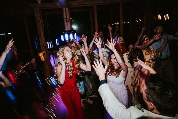 A lively reception dance floor scene with approximately 20 or more guests dancing with their arms raised in a darkened indoor venue with exposed wooden beams and posts. The space is lit with colorful DJ lighting in blue, red, and purple tones. In the foreground, a woman in a red jumpsuit and another in a lavender bridesmaid dress are prominently visible dancing and smiling, while a guest in a gold dress appears in the background. The groom, in a white shirt, is visible from behind in the lower right corner. Wide-angle shot capturing the energy of the full dance floor.