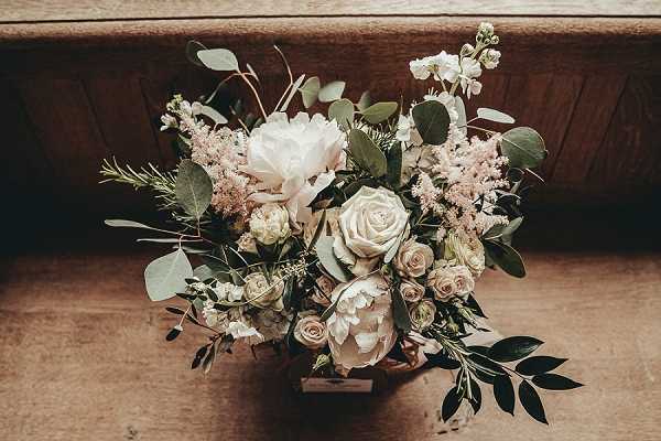 Close-up detail shot of a bridal bouquet resting on a wooden surface, likely a church pew or bench. The bouquet features large white peonies, blush and cream garden roses, soft pink astilbe, small white stock flowers, and abundant greenery including eucalyptus and ruscus foliage. The overall color palette is blush, cream, and soft white with deep green accents, styled in a loose, garden-gathered arrangement consistent with a romantic or boho aesthetic.
