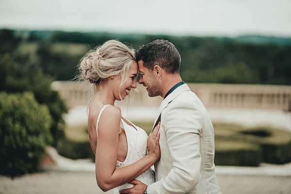 A close portrait of a bride and groom laughing together outdoors, their foreheads nearly touching in an candid, joyful moment. The bride wears a thin-strap lace or embroidered white gown with her blonde hair styled in a loose updo, while the groom wears a cream or off-white linen suit with a teal or dark green tie. The background is blurred but reveals a formal stone balustrade structure, suggesting a chateau or manor house setting, with manicured hedgerows visible in the foreground.