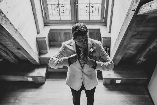 Black-and-white overhead portrait of a groom adjusting his jacket lapel or boutonniere while standing on a wooden staircase landing. The image is shot from directly above, creating a dramatic top-down composition that frames him between the wooden stair railings and exposed timber beams. He is wearing a light-toned blazer over a patterned shirt with dark trousers, and a small floral boutonniere is visible on his lapel. A leaded or decorative window in the background provides soft natural light, and the high contrast between the pale jacket and dark surroundings gives the image strong tonal depth.