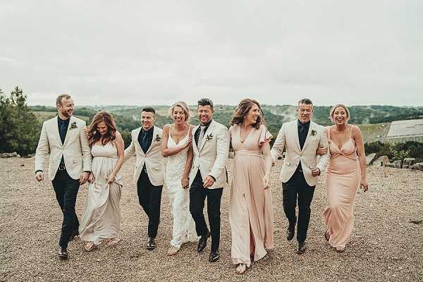 A bridal party of eight walks together across a gravel outdoor area, laughing and holding arms in a relaxed, candid group portrait. The bride wears a fitted lace white gown and the groom is dressed in a white blazer with dark trousers and a boutonniere. The two groomsmen wear navy suits with white blazers, while the three bridesmaids wear blush pink floor-length gowns in varying styles including a wrap silhouette and a draped spaghetti-strap dress. The overall styling theme is modern with a soft blush and navy palette. Wide shot taken at mid-distance with rolling countryside visible in the background.