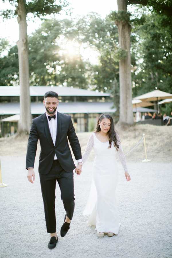 A couple walks hand-in-hand along a gravel path during what appears to be a post-ceremony or cocktail hour portrait moment. The groom wears a black tuxedo with a white dress shirt and black bow tie, paired with black loafers, and is smiling broadly. The bride wears a white long-sleeve A-line gown with lace sleeves and a simple scoop neckline, with her dark hair worn down in loose waves. In the background, a modern low-profile building with a flat roof and large glass windows is visible, along with beige market umbrellas and guests gathered in a shaded area. The overall styling is classic and minimal. Medium portrait shot with warm, soft backlit natural light filtering through the trees.