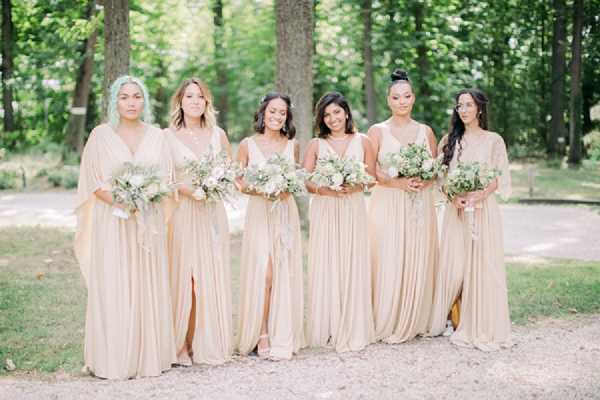 Six bridesmaids pose together outdoors on a gravel path in a wooded setting, walking and smiling toward the camera. All six wear floor-length champagne/nude gowns in varying styles — including a flowy wide-sleeve silhouette, spaghetti-strap with front slit, and a lace-detailed gown — creating a mix-and-match effect within a cohesive color palette. Each bridesmaid carries a bouquet of white and cream blooms, including what appear to be ranunculus and peonies, with soft greenery and trailing foliage in a loose, organic style. The shot is a mid-length portrait with a soft, airy aesthetic consistent with a boho or garden-style wedding.