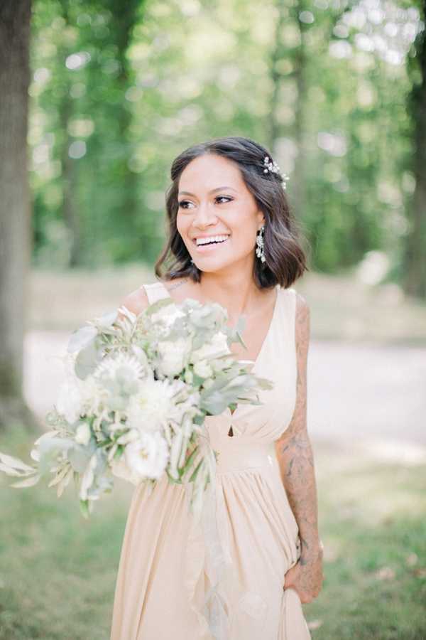 A portrait of a bridesmaid or bride standing outdoors in a wooded setting with a softly blurred background. She is wearing a champagne-gold sleeveless dress with a V-neckline and gathered waist, and has visible tattoos on both arms. Her hair is styled in a loose bob with a delicate crystal or pearl hair pin accessory, and she wears crystal drop earrings. She holds a loose, garden-style bouquet of white dahlias, white peonies, and soft green eucalyptus foliage. The shot is a medium portrait framing from the waist up, captured in a bright, airy style with natural light.