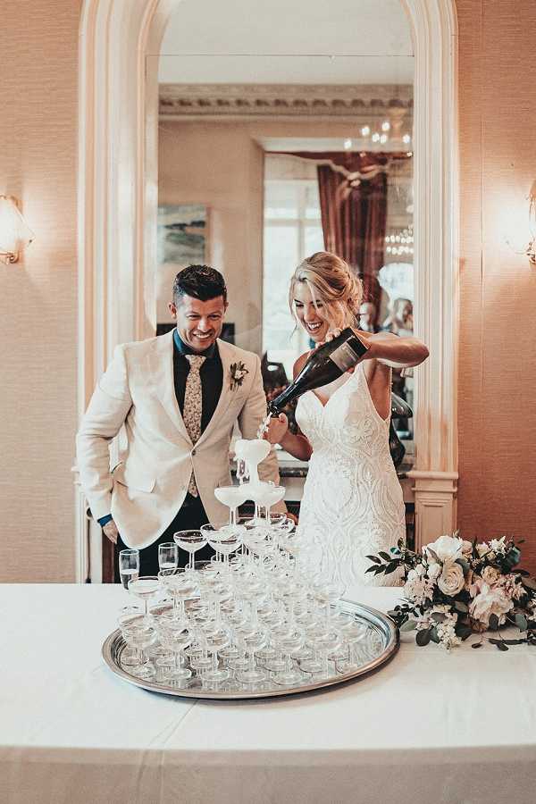 The bride and groom are pouring champagne over a coupe glass tower during their indoor reception, both laughing as champagne overflows down the pyramid. The groom wears a cream/off-white blazer with dark trousers, a floral tie, and a boutonniere, while the bride wears a fitted lace gown with a V-neckline. The setting is a classic interior room with warm blush-toned walls, wall sconces, a large arched mirror, and a chandelier visible in the reflection. A floral arrangement of blush and white blooms with eucalyptus greenery sits at the right end of the white-clothed table. Medium shot capturing both figures and the full glass tower on a silver tray.