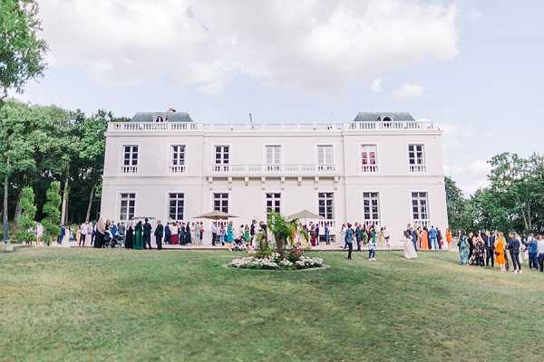 A wide exterior shot of a French chateau-style venue during what appears to be a cocktail hour or outdoor reception, with a large crowd of approximately 80–100 guests gathered on the terrace and lawn in front of the building. The venue is a two-story white neoclassical chateau with tall shuttered windows, balconies, and a mansard roof. Guests are dressed in colorful formal attire including jewel-toned dresses in emerald, orange, and blue. A small ornamental flower bed is visible in the foreground of the manicured lawn. Potential venue feature image.