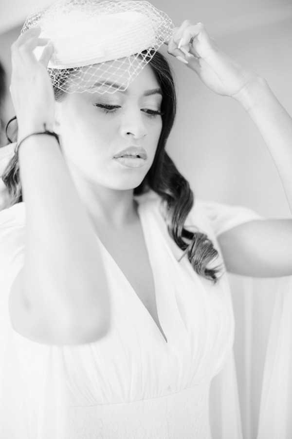 A black-and-white close-up portrait of a bride adjusting a birdcage veil and small pillbox-style hat on her head, eyes downcast. She is wearing a white gown with a deep V-neckline and flutter or cape sleeves, with her dark hair styled in loose waves. The image has soft, high-key tones with gentle contrast, giving it a light, airy feel consistent with a getting-ready moment. The background is plain and bright, keeping full focus on the bride and her accessories.