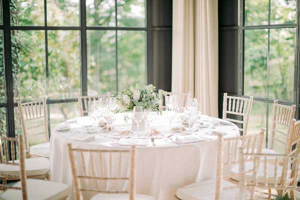 A close-to-medium shot of a round reception dining table set for approximately eight guests in a glass-walled conservatory or orangerie-style room with large black-framed windows overlooking garden greenery. The table is dressed in a white floor-length linen and set with crystal wine glasses, white folded napkins, and silver cutlery. The centerpiece is a low, compact arrangement of white flowers and green foliage in a white vessel. Seating consists of natural wood chiavari chairs with white cushions. The overall decor palette is white and ivory with greenery accents, reflecting a classic, clean aesthetic.