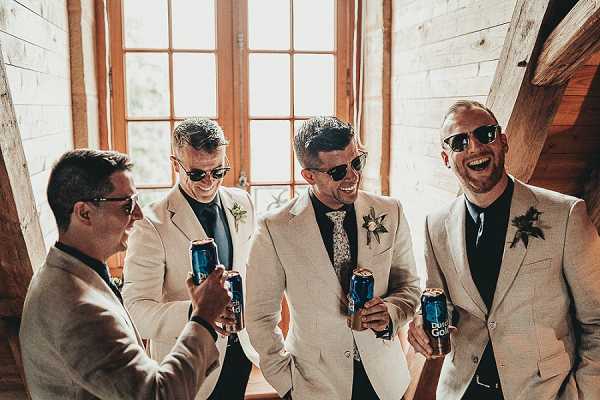 Four men — likely the groom and his groomsmen — are photographed getting ready indoors in what appears to be a rustic barn venue with exposed wooden walls and large wooden-framed windows. All four wear matching cream/off-white linen blazers with dark navy shirts and sunglasses, and each has a small floral boutonniere featuring greenery and small blooms. Three of the men are holding cans of Bud Gold beer and toasting, all laughing and smiling in a candid, relaxed moment. The shot is a medium group portrait with warm natural light coming through the windows behind them, and the styling has a casual, laid-back aesthetic within a rustic setting.