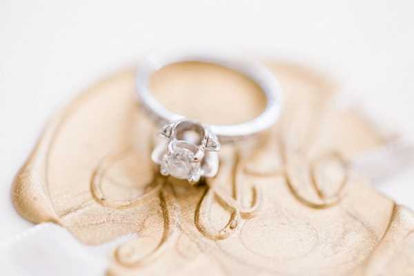 Close-up detail shot of a solitaire diamond engagement ring with a silver/platinum band, resting on a small gold-toned decorative ring dish with scrollwork detailing. The ring features a round-cut center stone set in a claw prong setting with small accent stones at the base. The background is softly blurred white, keeping full focus on the ring and dish.