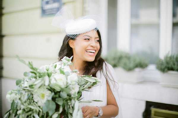A close-up portrait of a bride laughing and looking to one side, holding a loose bouquet of white flowers including what appear to be ranunculus and chrysanthemums with eucalyptus and greenery. She is wearing a white dress, a small white pillbox hat with a birdcage veil, and a silver watch. The setting appears to be outdoors on a Parisian street, with a light yellow building facade and a partially visible blue street sign in the background. The overall styling is classic French with a chic, minimal bridal aesthetic.