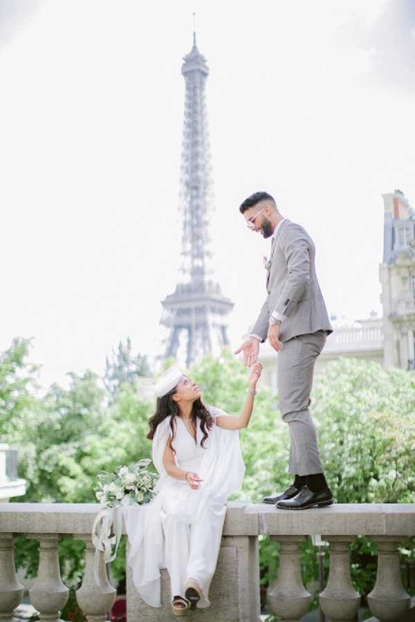 A couple portrait shot outdoors in Paris with the Eiffel Tower visible and slightly out of focus in the background. The bride is seated on a stone balustrade wearing a white wide-leg jumpsuit or flowing white trousers with a long white veil and wedge sandals, holding a loose bouquet of white blooms and greenery with trailing white ribbon. The groom stands on the balustrade beside her wearing a light gray suit with black dress shoes and a small white boutonniere, reaching down to hold her hand as she looks up at him. The composition is a full-length portrait with a bright, slightly overexposed airy quality, and the Haussmann-style architecture of Paris is partially visible to the right.