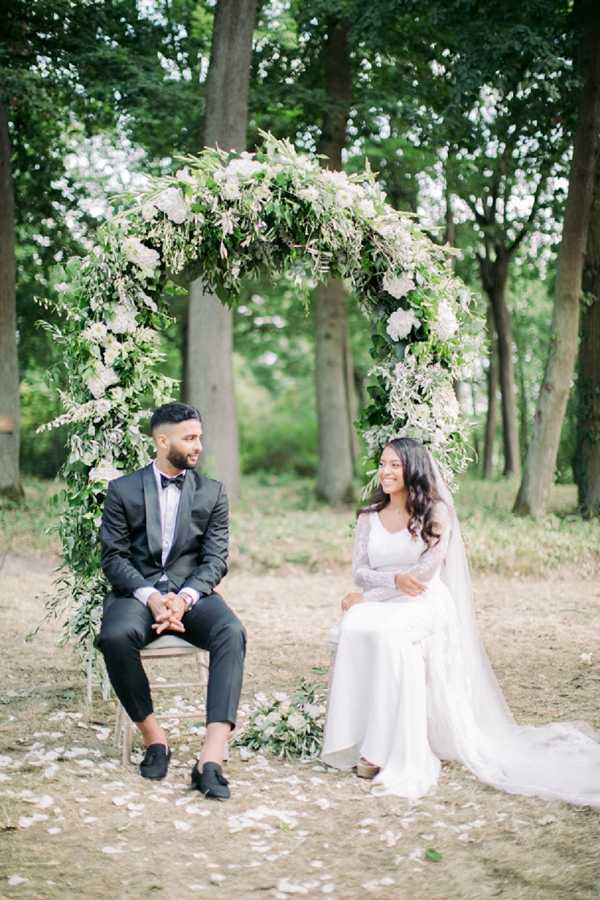 A couple sits on chairs beneath a large circular floral arch during what appears to be an outdoor ceremony or portrait session set within a wooded area. The arch is densely covered with lush green foliage, white hydrangeas, and trailing greenery. The groom wears a dark charcoal suit with a black bow tie and black loafers, while the bride wears a long-sleeved ivory lace gown with a flowing veil and has her dark hair worn loose in waves. A small white floral bouquet rests on the ground near her feet, and white flower petals are scattered across the dirt ground around them. The couple is seated facing each other and smiling, giving the image a relaxed, candid quality. The overall styling leans toward a classic garden aesthetic with a green and white color palette. This is a medium-distance portrait shot.