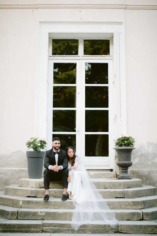 A couple portrait taken outdoors on the stone steps of what appears to be a French chateau or manor house. The groom is dressed in a black tuxedo with a bow tie and black loafers, while the bride wears a white long-sleeved gown with a long flowing veil that cascades down the steps. Both are seated facing slightly away from each other, looking in different directions rather than at the camera. Two dark planters with pale pink hydrangeas flank the white-framed French door behind them. The overall styling is classic and minimal, with a white and black color palette. Medium-distance portrait shot with the architectural facade serving as a centered backdrop.