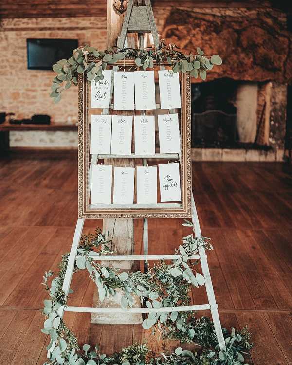 A close-up detail shot of a wedding seating chart display set up indoors in what appears to be a rustic stone-walled venue with a large fireplace and warm wood flooring visible in the background. The seating plan cards are printed in black script on white card stock and are clipped to horizontal rungs inside an ornate gold-framed display mounted on a whitewashed wooden ladder-style easel. The text on the cards includes 'Please find your seat' and 'Bon appétit' among the table name headers. Eucalyptus garlands with round silver-green leaves are draped around the top of the easel and pooled loosely at its base, giving the display a natural, greenery-forward rustic aesthetic with minimal floral color.