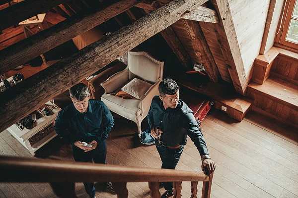 Getting-ready scene captured from above, showing two men ascending a wooden staircase inside a rustic alpine chalet with exposed heavy timber beams and warm wood plank flooring. Both men are dressed in dark navy shirts with dark trousers — one holds a phone, the other holds a drink. A cream upholstered armchair with an orange cushion is visible in the background loft area. The overhead angle and warm wood tones give the shot a candid, documentary feel.