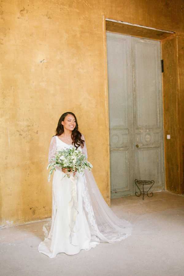 A bridal portrait taken indoors, with the bride standing against a warm ochre-yellow plastered wall beside a weathered sage-green double door. The bride wears a long-sleeve lace wedding dress with a flowing train and a long tulle veil, and holds a loosely arranged bouquet of white blooms, eucalyptus, and trailing greenery with ivory ribbon streamers. Her dark hair falls in loose waves over her shoulders and she is smiling softly downward. The composition is a full-length portrait with a classic, understated romantic styling that suits the rustic French chateau interior aesthetic.